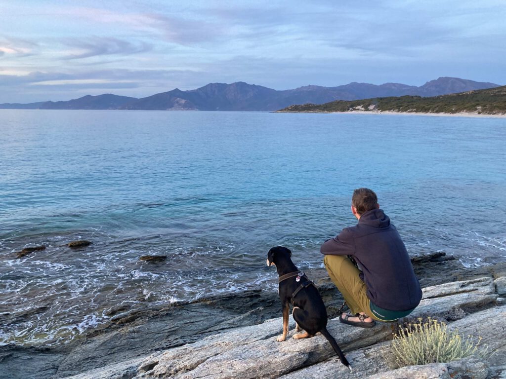 Mathias mit seinem Hund blickt über einen See mit Bergen im Hintergrund