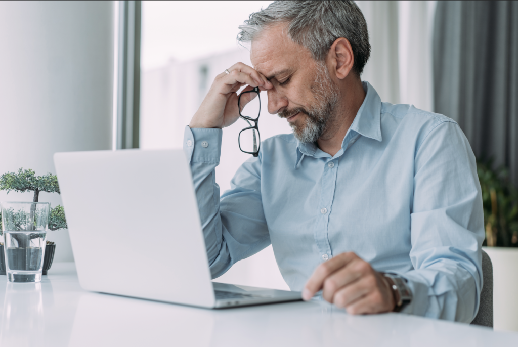 Businessmann ist gestresst im Büro vor dem Computer, hält die Brille in der Hand und reibt sich die Augen